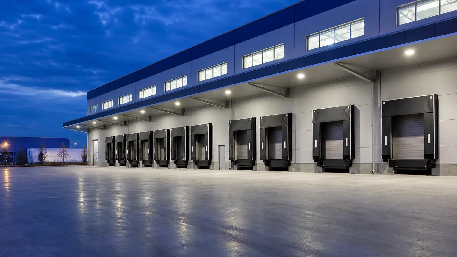 Modern logistics warehouse exterior at twilight with clean loading-dock canopy and empty truck court